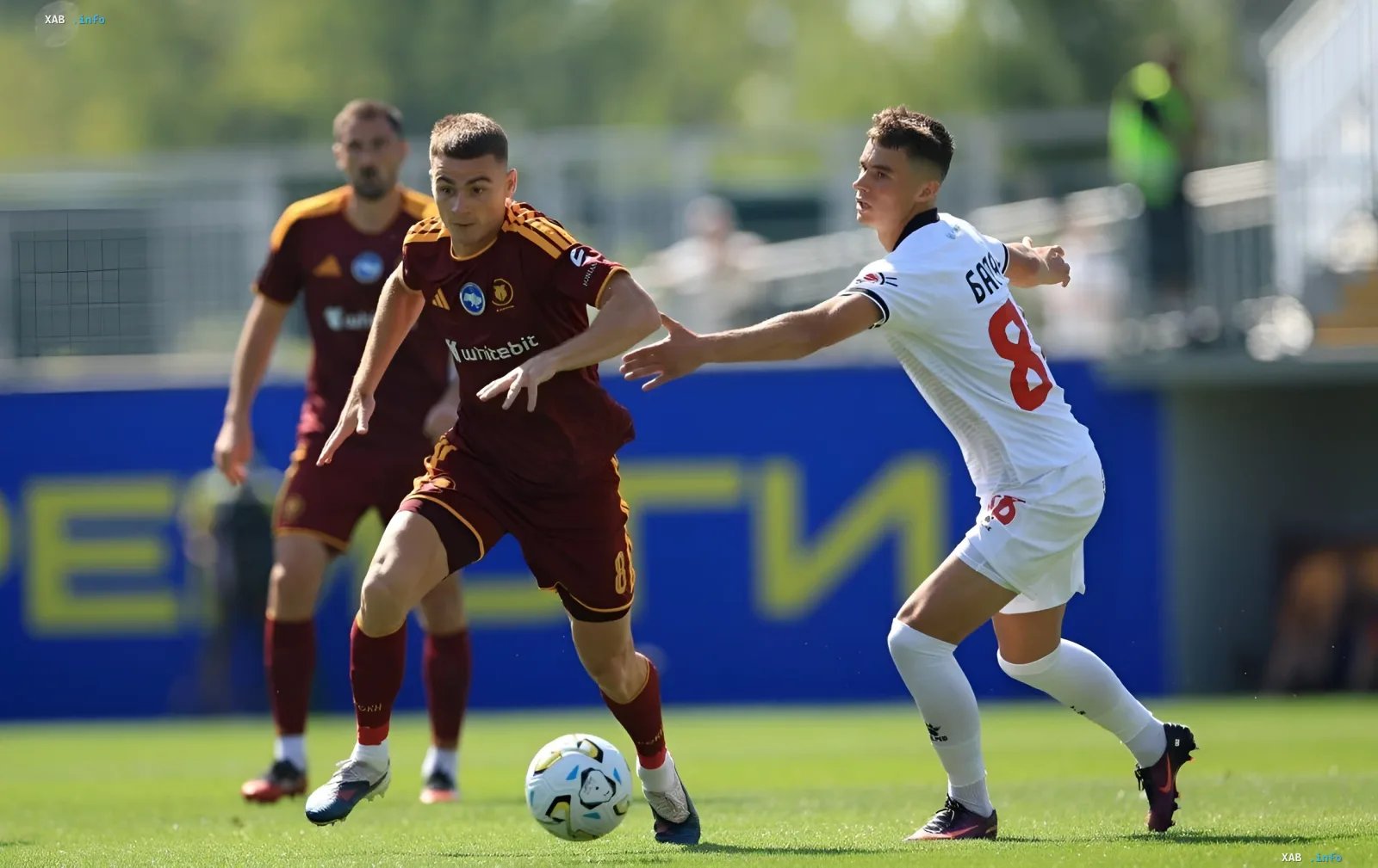 Ein Spieler von 'Charkiw' in einem kastanienbraunen Trikot dribbelt den Ball unter Druck eines 'Veres'-Verteidigers in Weiß, was die intensive Auseinandersetzung im Spiel illustriert