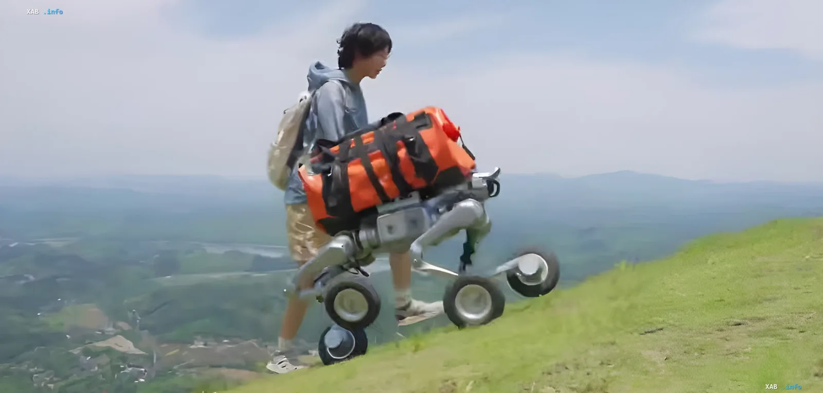 Person using Unitree AS2-W robot to haul a heavy orange backpack up a steep grassy slope, demonstrating the platform's actual payload capacity.