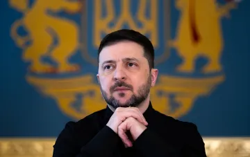Президент Украины Владимир Зеленский задумчиво смотрит в сторону, на фоне герба Украины, обсуждая причины прорыва баллистической ракеты в Киев