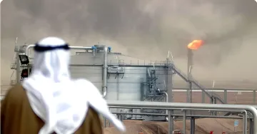 Рабочий в традиционной арабской одежде наблюдает за нефтеперерабатывающим заводом с горящим факелом на фоне пыльной атмосферы — символ уязвимости энергетической инфраструктуры Ближнего Востока перед угрозой удара