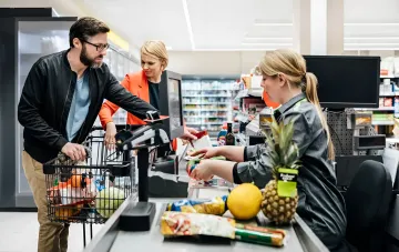 Einkäufer in einem 'Nahkauf'-Supermarkt in der Ukraine: Mann und Frau bezahlen an der Kasse, was die wachsende Beliebtheit lokaler Einzelhandelsformate während des Krieges veranschaulicht