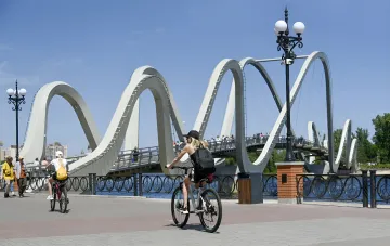 Велосипедисты на набережной в солнечный день на фоне арочного моста — иллюстрация к прогнозу погоды на 2 августа с жарой и грозами