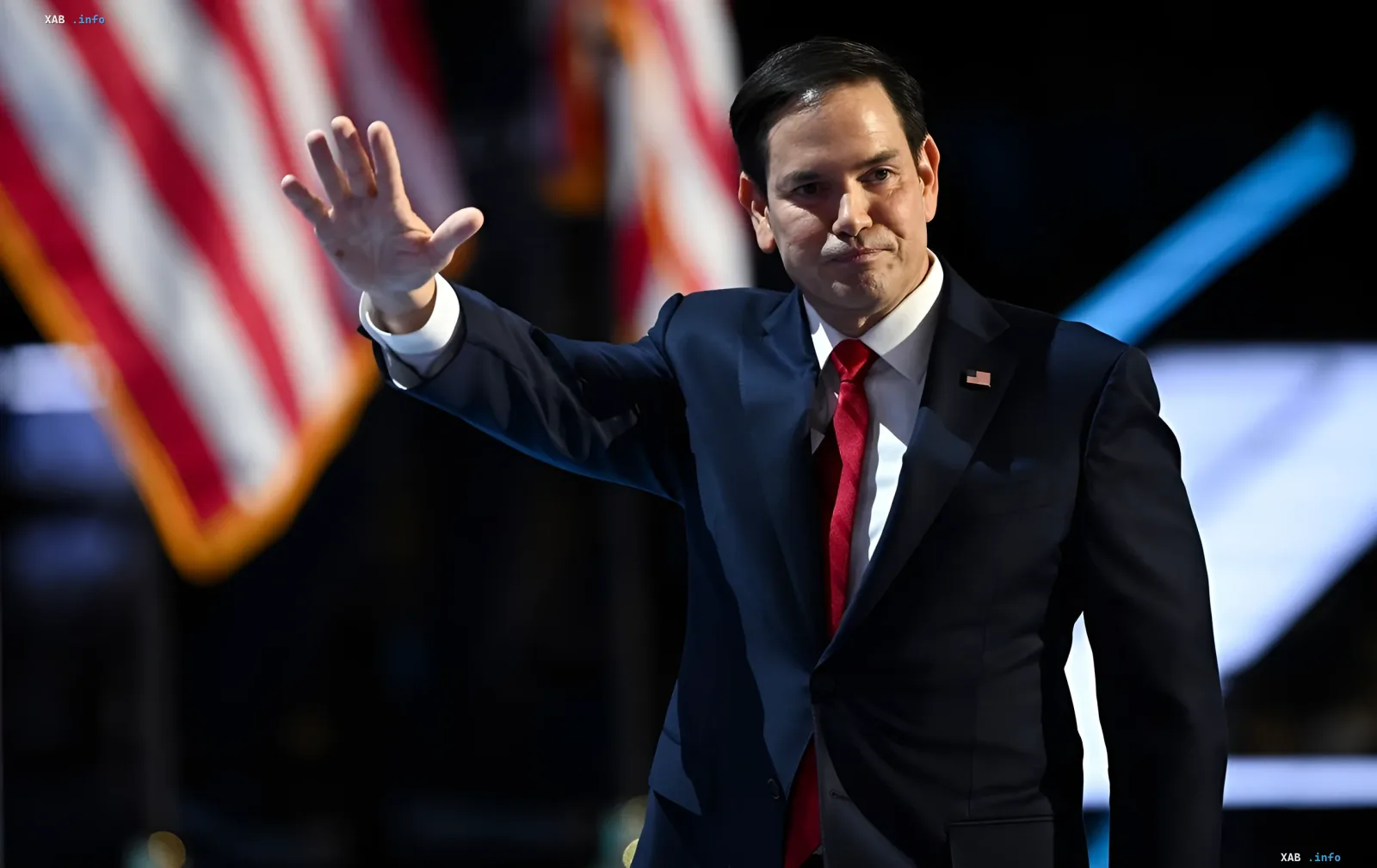 Marco Rubio in a blue suit and red tie waves his hand against the backdrop of the American flag, symbolizing the US readiness for peace negotiations