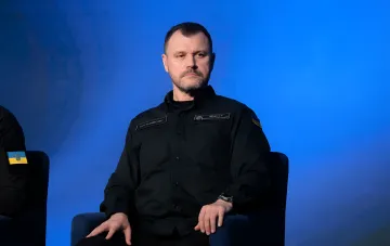 Игорь Клименко в черной форме с украинским флагом на фоне синего экрана, символизирующий его новую роль в СНБО