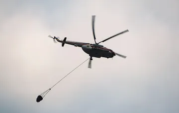 Firefighting helicopter in flight with a suspended water bucket, symbolizing the bravery of rescuers who died in the Athens tragedy