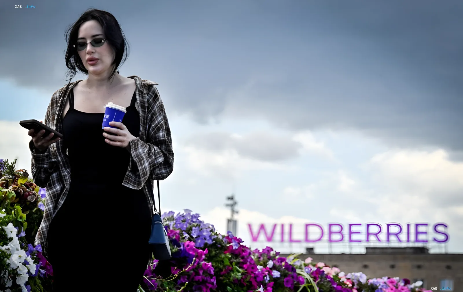 Woman in sunglasses holding a smartphone and coffee in front of the Wildberries logo, illustrating new safety rules for employees