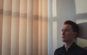 Мужчина задумчиво смотрит в сторону, освещённый полосами света от жалюзи — метафора внутреннего конфликта при распознавании манипуляции в общении