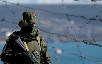Российский военный в камуфляже стоит у колючей проволоки на фоне моря, символизируя напряженную обстановку в Крыму после инцидента с гибелью мирных жителей.