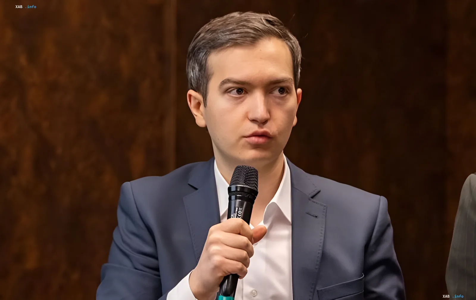 Young man in business suit holding microphone against wooden panel background — expert discusses risks of maritime corridor shutdown for metallurgy in 2026