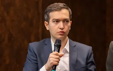 Молодий чоловік у діловому костюмі з мікрофоном на тлі дерев'яної панелі — експерт обговорює ризики зупинки морського коридору для металургії у 2026 році