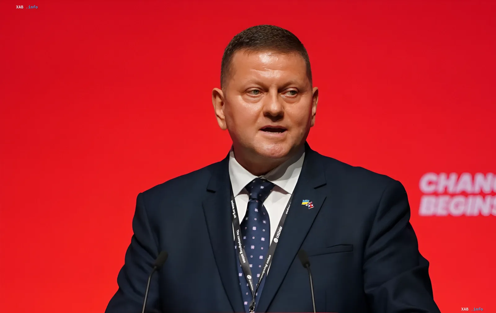 Президент Украины Владимир Зеленский выступает с речью на фоне красного баннера с надписью 'CHANGE BEGINS', что символизирует политические реформы и диалог с НАТО