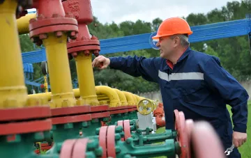 Работник в каске и защитной форме управляет газовыми трубами и клапанами на газопроводе, символизируя контроль над транспортировкой и накоплением газа в Украине.