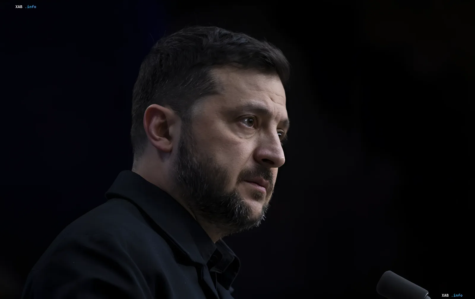 Президент Украины Владимир Зеленский в черной рубашке на фоне темного фона, выражает серьезность и решимость, осуждая атаку дрона на овощника в Херсоне