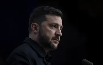 Президент Украины Владимир Зеленский в черной рубашке на фоне темного фона, выражает серьезность и решимость, осуждая атаку дрона на овощника в Херсоне