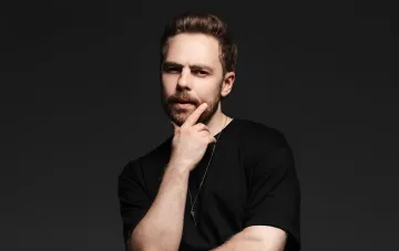 Anton Timoshenko in a black t-shirt thoughtfully touching his chin against a dark background — illustration for an interview about money, envy, and war