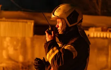 Пожежник у захисному шоломі та формі з відбиваючими смугами тримає радіостанцію на тлі вогняного світла вночі, ймовірно, під час ліквідації наслідків атаки в Києві