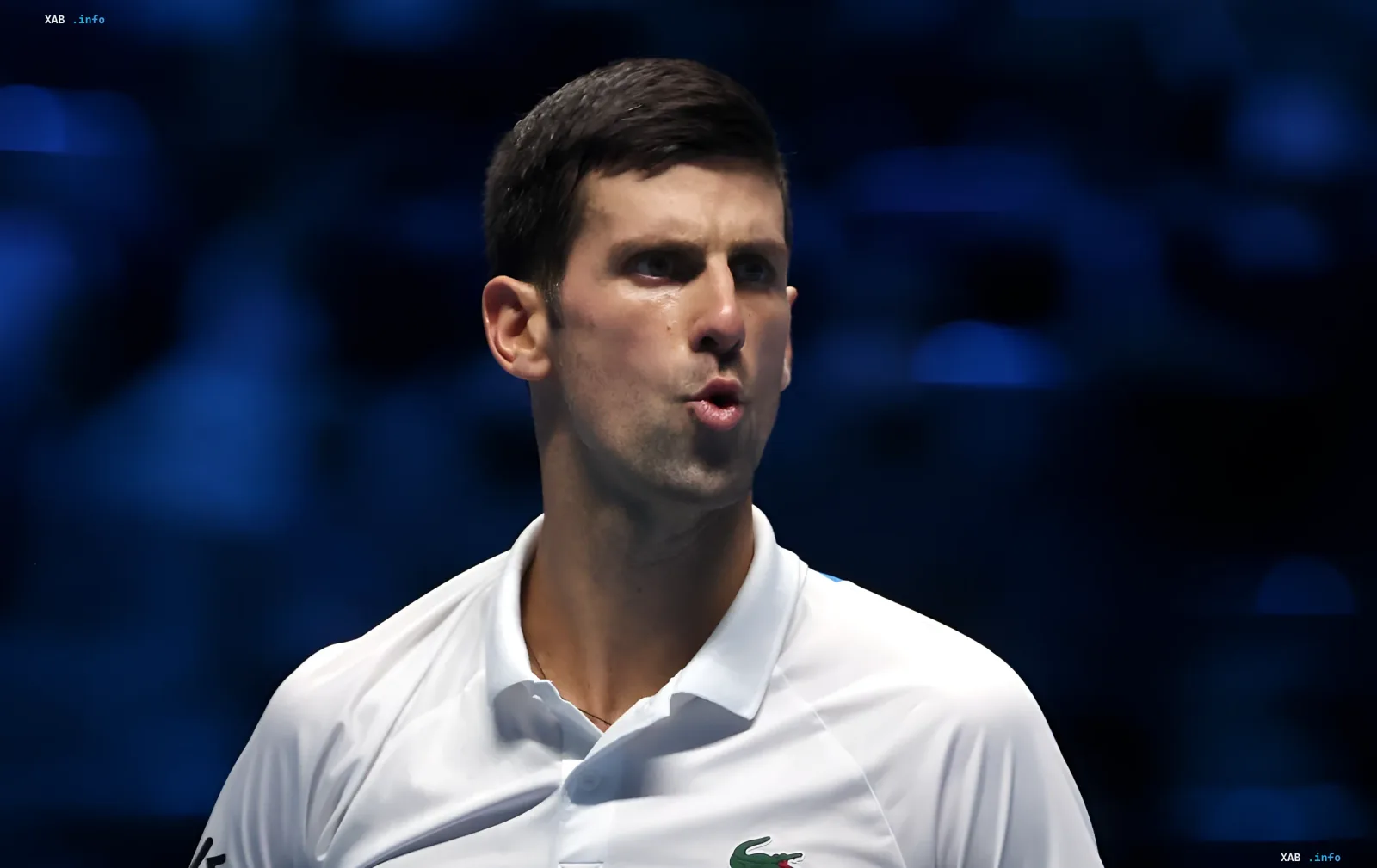 Новак Джокович в белой теннисной форме с логотипом Lacoste, задумчиво смотрит в сторону на фоне синего стадиона — символ его призыва к реформам в теннисе