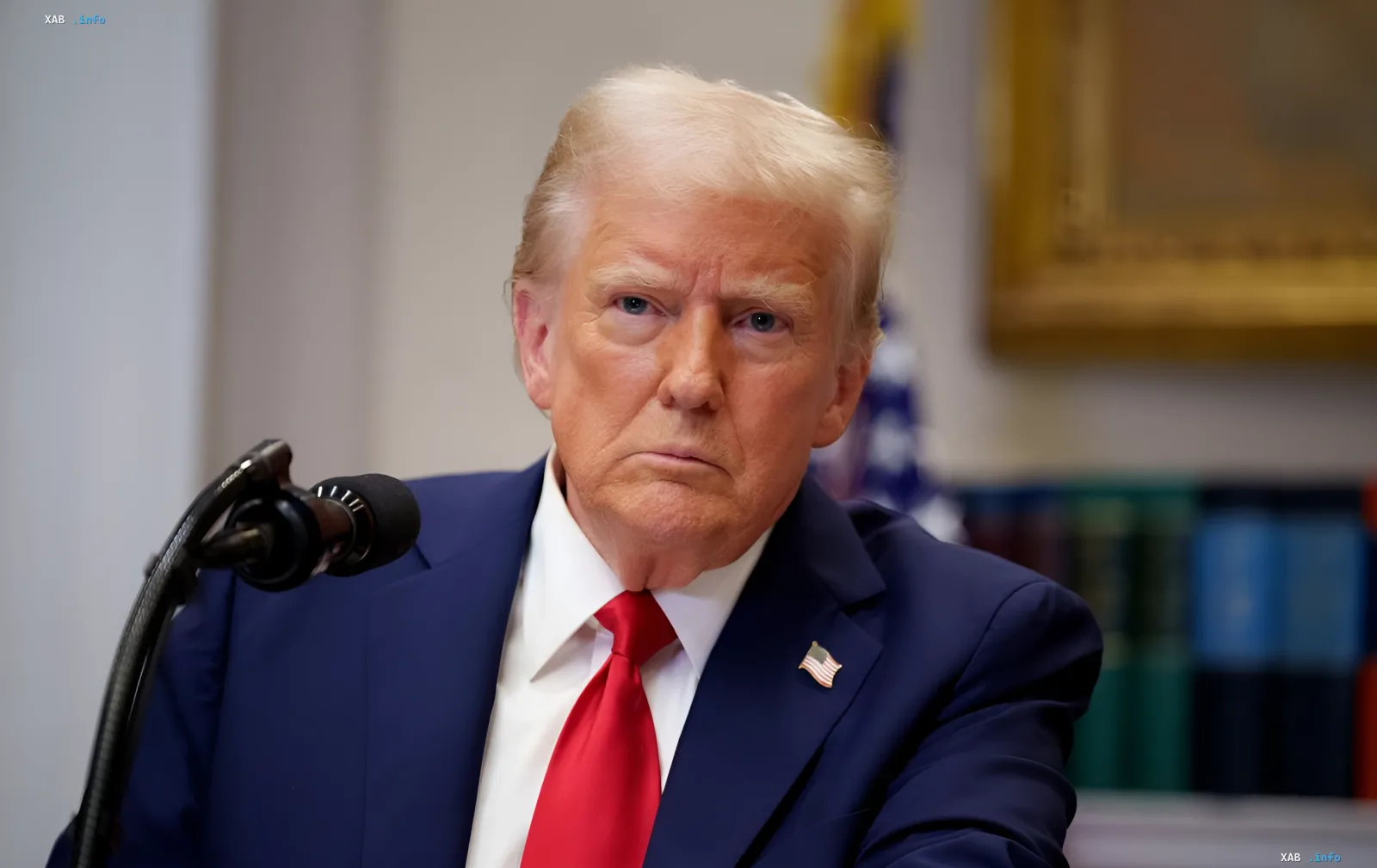 Дональд Трамп в синем костюме и красном галстуке с американским флагом на лацкане, сидящий за столом с микрофоном — символ переговоров по Ормузскому проливу