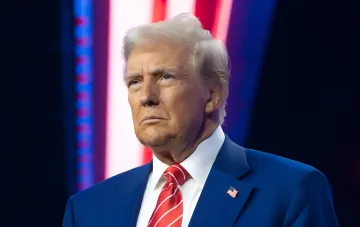 Дональд Трамп у синьому костюмі з американським прапором на лацкані, на тлі червоно-синіх світлових смуг — ілюстрація до новини про його заяву щодо підготовки атаки на Іран з часів Другої світової війни