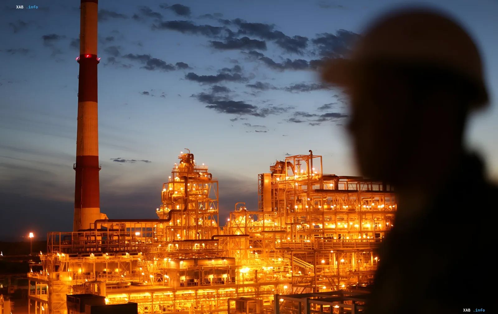 Ночной вид нефтеперерабатывающего завода с горящими огнями и высокой трубой, символизирующий снижение переработки нефти в России до уровня 2002 года после ударов