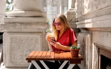 Woman in red blouse sits on a sunny terrace sipping a drink while using her smartphone — illustrating the risk of device overheating in extreme heat