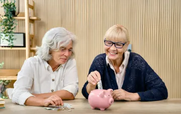 Zwei ältere Frauen zählen Geld und legen einen Schein in ein Sparschwein, was das Thema Rentenleistungen und finanzielle Sicherheit illustriert