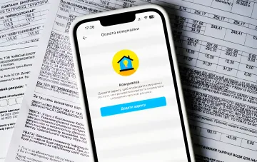 Смартфон с приложением для оплаты коммунальных услуг лежит на фоне счетов за ЖКХ, символизируя проблему долгов за уничтоженное жилье в Украине