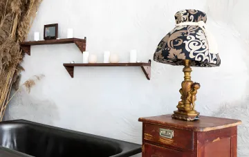 Vintage Bronzelampe mit Empire-Schirm auf einem antiken Holzschrank in einem Raum mit weißer strukturierten Wänden und Regalen mit Kerzen