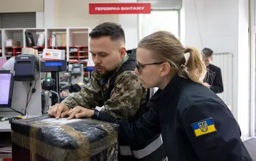 Волонтер у камуфляжі та працівник пошти з українським прапором на рукаві перевіряють посилку — ілюстрація до теми податкових пасток для волонтерів