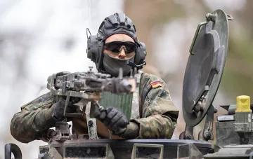 Солдат в немецкой камуфляжной форме Flecktarn с флагом Германии на рукаве управляет пулеметом на бронетехнике