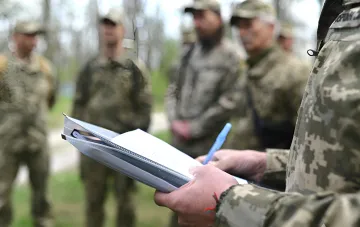 Сотрудник ТЦК заполняет документы в присутствии группы мужчин в военной форме на фоне леса, иллюстрируя тему жалоб на жестокость призывников в Одессе