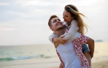 Young couple on the beach: man carries woman on his back, both smiling — illustration for article about idealizing partners in love by zodiac signs