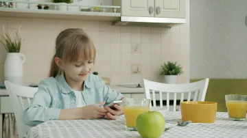 Mädchen beim Frühstück schaut auf Smartphone, illustriert digitale Kontrolle und SMS-Verbot für Kinder