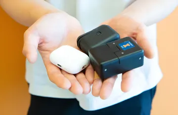 Größenvergleich: weißes AirPods-Etui und zwei schwarze Prototypen eines Molekularanalysators für die häusliche Krebsdiagnostik, die die Kompaktheit des neuen Geräts zeigen