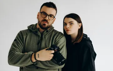 Photographers Liberovs with Sony camera against white studio background — man in glasses and hoodie holds camera, woman in black jacket beside him