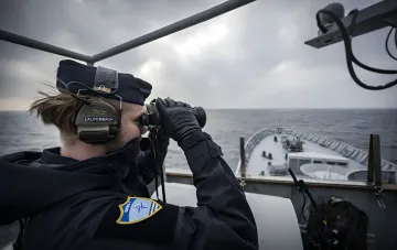 Моряк ВМС Германии с опознавательным знаком НАТО наблюдает за горизонтом в бинокль с палубы корабля