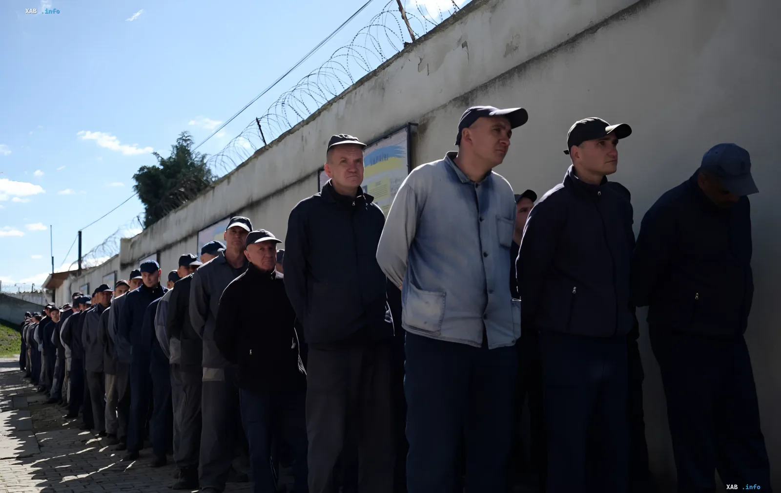 Украинские военнопленные в российской тюрьме стоят в очереди у стены с колючей проволокой, иллюстрируя реальность вместо пропагандистских обещаний «улучшения» условий содержания.