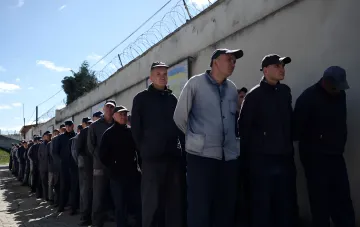 Украинские военнопленные в российской тюрьме стоят в очереди у стены с колючей проволокой, иллюстрируя реальность вместо пропагандистских обещаний «улучшения» условий содержания.