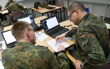 Немецкие военнослужащие в камуфляжной форме работают за ноутбуками в учебном классе, анализируя данные для превентивной борьбы с угрозами.