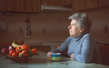 Пенсионерка сидит за столом с чашкой чая, задумчиво глядя в окно — иллюстрация к статье о получении субсидии при прописанных, но не живущих детях