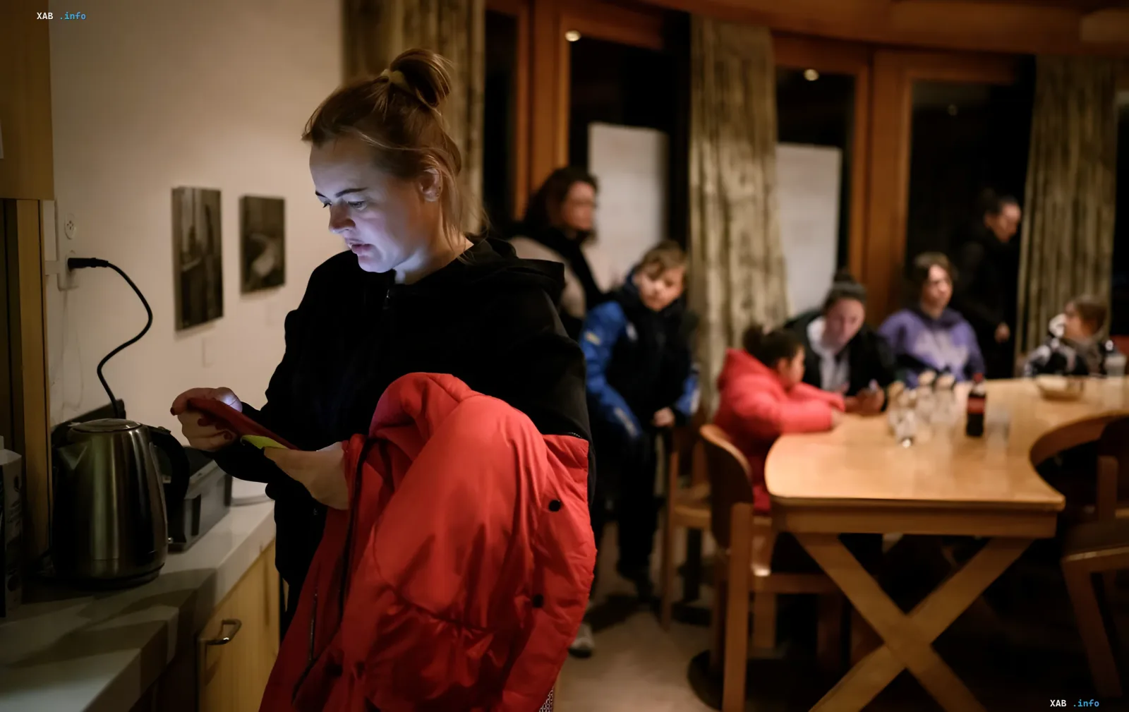 Woman in red jacket checking phone in kitchen while others sit at table — visual for Zurich tightening social aid rules for Ukrainians with S status