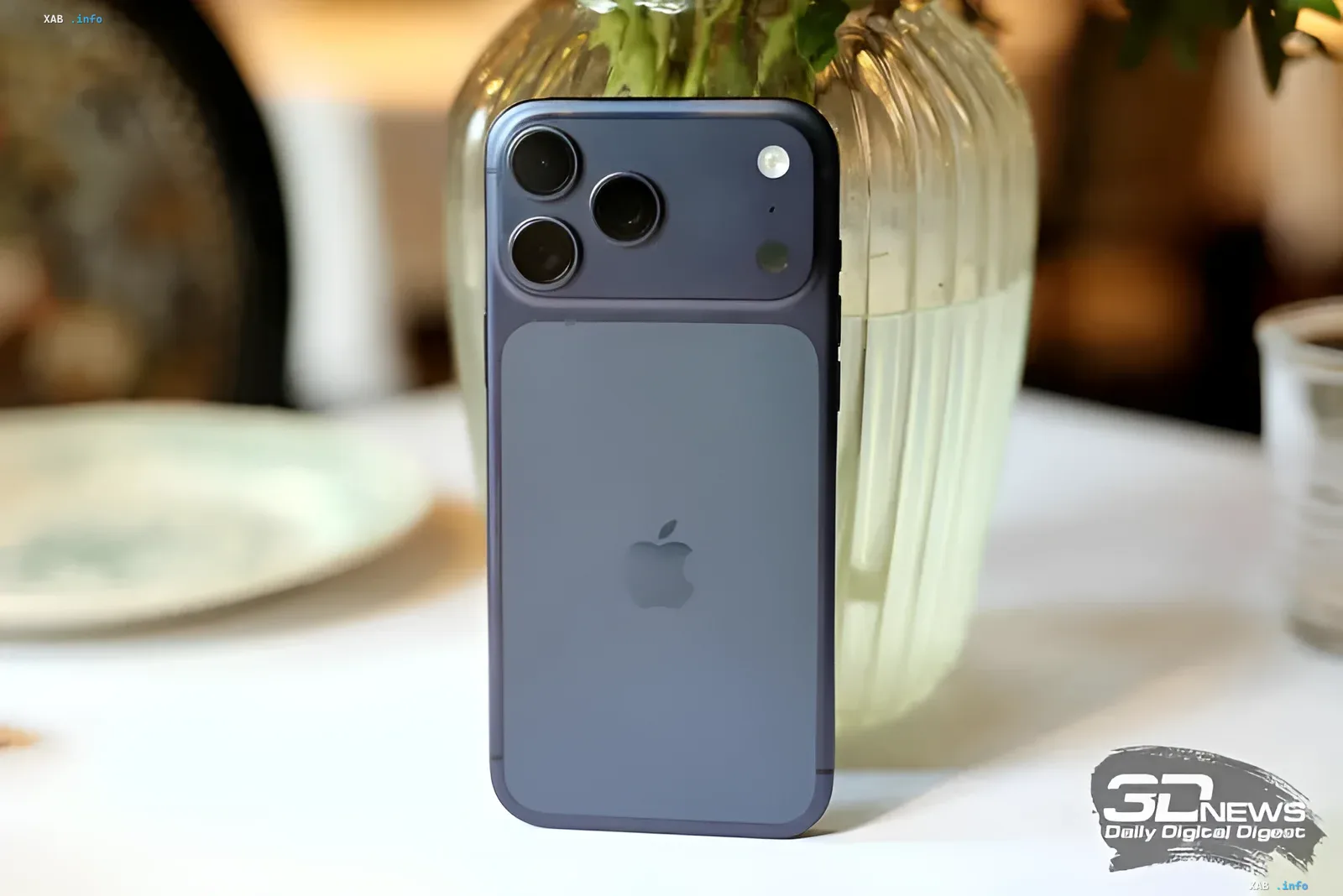 Синий iPhone с тремя камерами и логотипом Apple на фоне стеклянной вазы, иллюстрирующий тему защиты от дипфейков в iOS 27