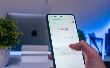 Smartphone mit geöffnetem Google-Startseite, im Hintergrund ein moderner Arbeitsplatz; Illustration zur Nachricht über Forderung französischer Medien nach Entschädigung von Google wegen KI-Zusammenfassungen und 38% Traffic-Rückgang