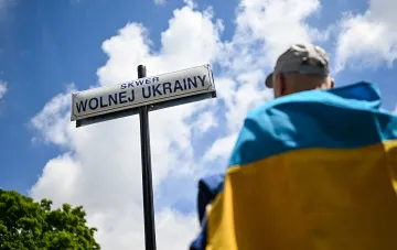 Человек с украинским флагом на фоне знака 'Площадь Свободной Украины' в Польше, иллюстрирующая тему ксенофобии и поддержки депортации безработных украинцев.