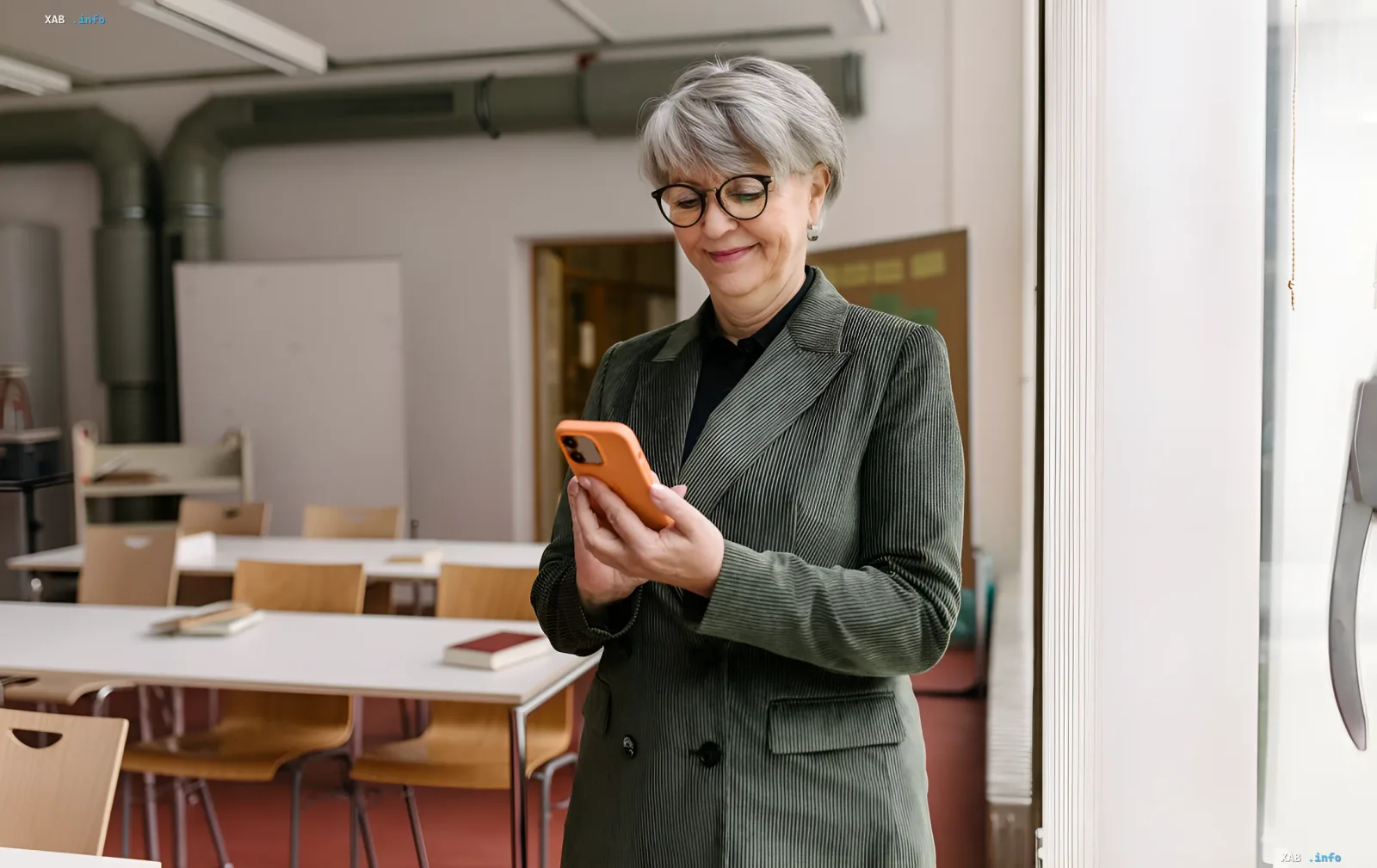 Rentnerin mit grauen Haaren, Brille und grünem Blazer lächelt, während sie auf ein Smartphone mit orangefarbenem Case in einem Klassenzimmer schaut – Illustration zugänglicher Konnektivität für Senioren in der Ukraine im Jahr 2026