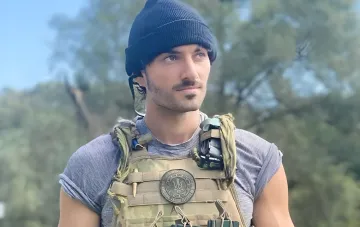 Connor Kennedy in military gear with a Ukrainian symbol on his chest, against a forest backdrop
