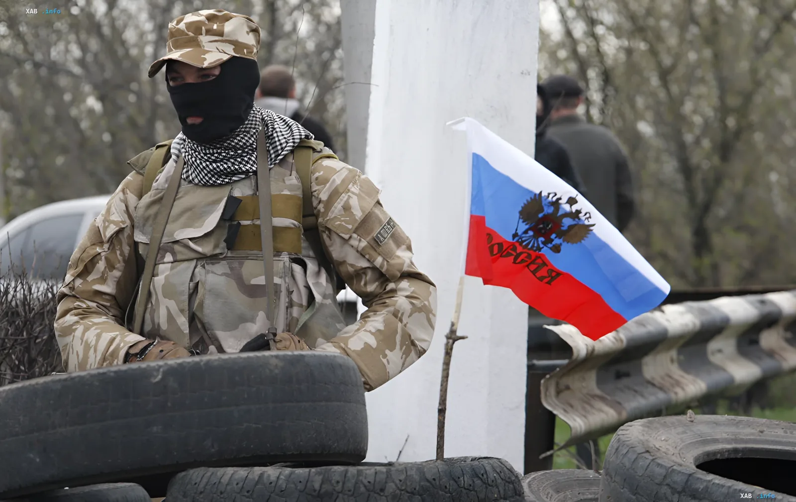 Военный в камуфляже с российским флагом на фоне блокпоста из шин — иллюстрация к новости об отмене приграничного контроля ФСБ