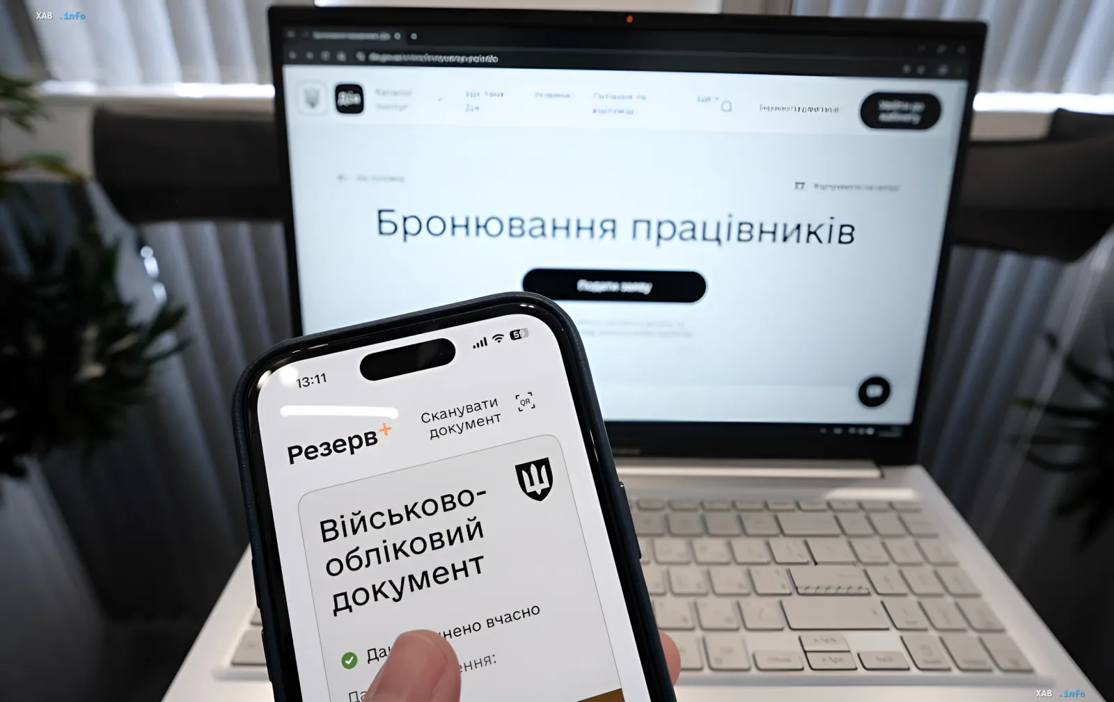 Смартфон с приложением «Резерв+» показывает военно-учетный документ, на фоне — ноутбук с порталом бронирования работников в Украине
