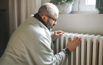 Mann unter einer Decke wärmt seine Hände an einem Heizkörper, was die Bedeutung der Unterstützung des Energiesystems der Ukraine vor dem Winter symbolisiert