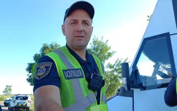 Украинский полицейский в жилете с надписью 'ПОЛІЦІЯ' и нашивкой 'Херсонская область' стоит у патрульного автомобиля на дороге, символизируя остановку эвакуации из Херсона